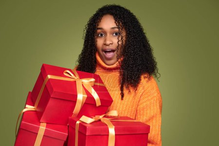 Foto de Surprised African American female with curly dark hair and mouth opened carrying red present boxes with golden ribbons against green background - Imagen libre de derechos