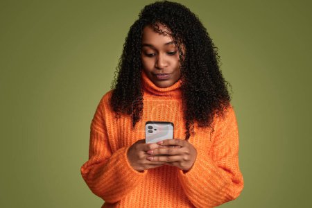 Photo for Ethnic trendy teenage young woman in bright orange sweater using smartphone, messaging friends, on green color background - Royalty Free Image