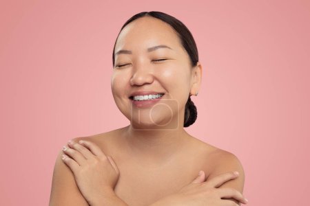 Téléchargez les photos : Heureuse jeune femme ethnique au visage propre et sans maquillage touchant les épaules nues tout en se tenant les yeux fermés sur fond rose - en image libre de droit