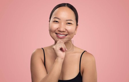 Foto de Mujer étnica positiva con una piel suave perfecta y hombros desnudos sonriendo mientras mira a la cámara contra el fondo rosa - Imagen libre de derechos