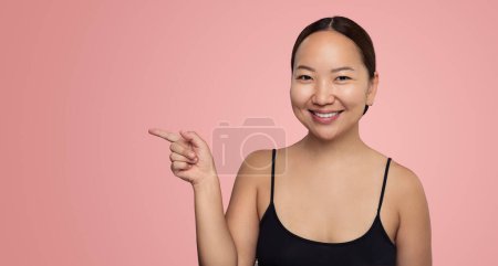 Photo for Cheerful young Asian female model with dark hair and perfect skin in black top, smiling and looking at camera while pointing aside against pink background - Royalty Free Image