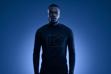 Téléchargez les photos : Auto assuré jeune athlète homme afro-américain barbu aux cheveux foncés en vêtements de sport noirs debout sur fond bleu et en regardant la caméra - en image libre de droit