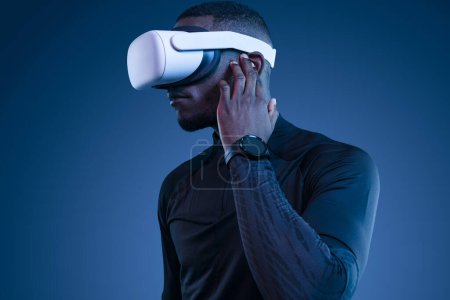 Foto de Joven afroamericano concentrado en ropa elegante y reloj inteligente ajustando auriculares TWS y viendo video en gafas VR modernas contra fondo azul - Imagen libre de derechos