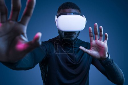 Téléchargez les photos : Jeune homme afro-américain concentré portant des lunettes VR et des vêtements de sport gesticulant tout en touchant un écran invisible et explorant le cyberespace sur fond bleu - en image libre de droit