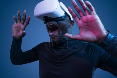 Téléchargez les photos : Jeune Afro-Américain effrayé en vêtements noirs criant émotionnellement tout en explorant la réalité virtuelle dans un casque moderne sur fond bleu - en image libre de droit