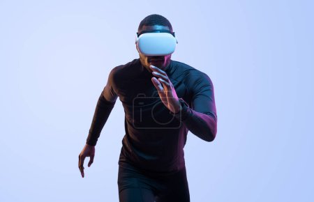 Téléchargez les photos : Athlète musclé afro-américain déterminé en vêtements de sport noirs en casque de réalité virtuelle blanche fonctionnant sur fond bleu - en image libre de droit