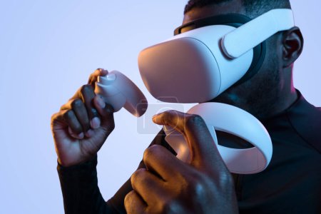 Photo for Crop young African American male millennial pushing buttons of controller while playing video game in VR goggles in purple neon studio - Royalty Free Image