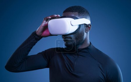 Foto de Hombre negro joven concentrado con cabello oscuro y barba en ropa deportiva ajustando gafas VR modernas mientras ve video en estudio azul - Imagen libre de derechos