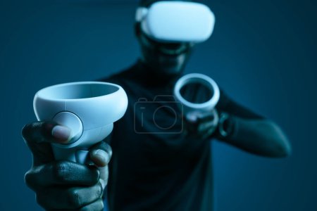 Photo for Crop anonymous African American male gamer in white virtual reality headset holding controllers while playing videogame in dark room - Royalty Free Image