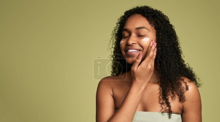 Téléchargez les photos : Contenu jeune femme afro-américaine aux longs cheveux bouclés foncés et aux épaules nues, souriant les yeux fermés tout en appliquant de la crème hydratante sur le visage après la douche sur fond vert - en image libre de droit
