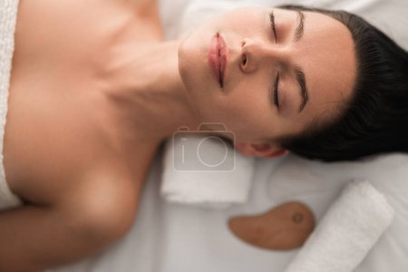 Téléchargez les photos : Vue de dessus de jeune femme calme enveloppée dans une serviette couchée sur un canapé avec les yeux fermés après un massage relaxant avec Gua Sha - en image libre de droit