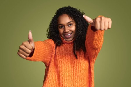 Photo for Young carefree ethnic woman wearing vivid orange sweater showing thumbs up and smiling on green background - Royalty Free Image