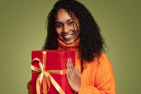 Foto de Joven hembra negra positiva con pelo rizado en suéter de punto sonriendo y mirando a la cámara mientras muestra la caja de regalo roja de cumpleaños con fondo verde - Imagen libre de derechos