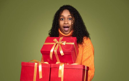 Foto de Mujer afroamericana sorprendida con el pelo rizado en suéter naranja sosteniendo cajas de regalo sobre fondo verde - Imagen libre de derechos