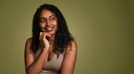 Téléchargez les photos : Charmante jeune femme afro-américaine avec les cheveux bouclés toucher le menton et regarder vers le haut rêveur sur fond vert - en image libre de droit