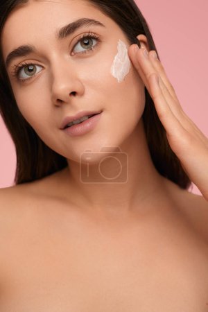 Photo for Crop young female with bare shoulders and applying moisturizing cream on face, while looking at camera on pink background - Royalty Free Image