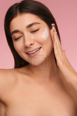 Photo for Positive young female model with closed eyes and bare shoulders applying moisturizing cream on face during skincare routine against pink background - Royalty Free Image