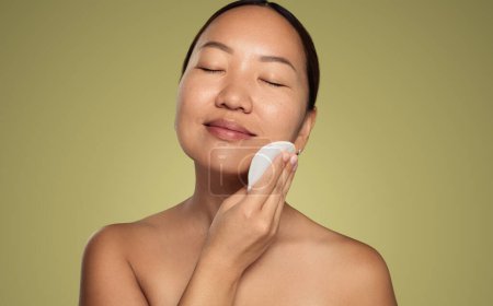 Téléchargez les photos : Jeune femme asiatique aux épaules nues appliquant du toner et essuyant le visage avec du coton tout en gardant les yeux fermés sur fond vert - en image libre de droit