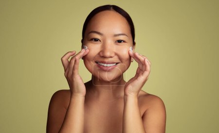 Téléchargez les photos : Joyeux jeune modèle féminin asiatique aux cheveux foncés et aux épaules nues souriant et regardant la caméra, tout en appliquant de la crème hydratante sur le visage pendant la routine de soins de la peau sur fond vert - en image libre de droit