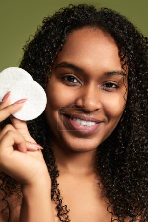 Photo for Cheerful young black female with curly dark hair showing white soft cotton pads looking at camera against green background - Royalty Free Image