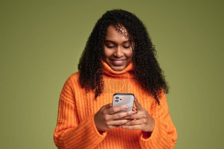 Téléchargez les photos : Femme afro-américaine moderne positive avec des cheveux bouclés en pull orange naviguant sur les médias sociaux sur un téléphone mobile tout en se tenant debout sur fond vert - en image libre de droit