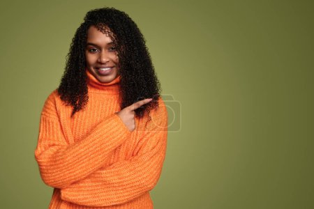 Photo for Confident young black female model with dark Afro hair in warm orange sweater smiling and looking at camera while pointing aside against green background - Royalty Free Image