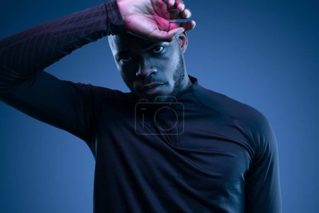 Foto de Agotado joven atleta afroamericano sin afeitar en ropa deportiva tocando la frente y mirando a la cámara contra el fondo azul - Imagen libre de derechos