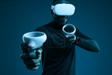 Foto de Cultivo positivo joven negro hombre en ropa casual sonriendo y pulsando botones de VR joysticks mientras juega videojuego en auriculares contra fondo azul - Imagen libre de derechos