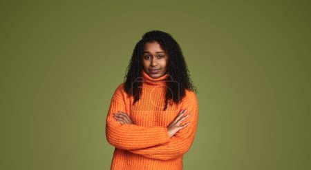 Foto de Joven mujer afroamericana con el pelo rizado en suéter naranja cruzando las manos mientras está de pie sobre fondo verde mirando a la cámara - Imagen libre de derechos