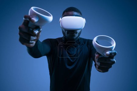 Téléchargez les photos : Jeune joueur afro-américain concentré à col roulé noir aux cheveux foncés, utilisant des contrôleurs VR tout en jouant à un jeu vidéo dans un casque moderne sur fond bleu - en image libre de droit