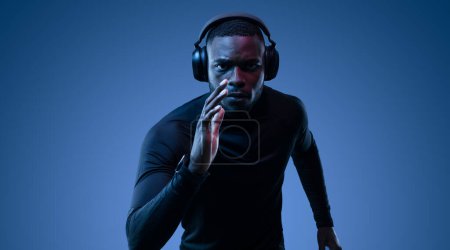 Photo for Determined African American sportsman in black turtleneck and earphones running on blue background in dark room while listening to music and looking at camera - Royalty Free Image