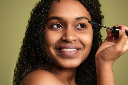 Téléchargez les photos : Headshot de charmante jeune femme noire avec des cheveux bouclés appliquant mascara sur les cils tout en souriant et en regardant loin sur fond vert - en image libre de droit