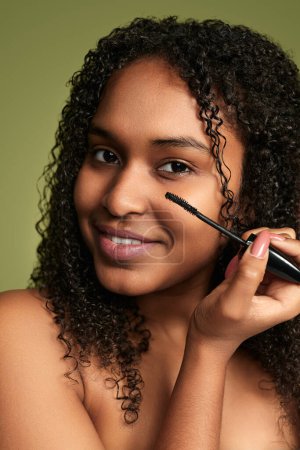 Téléchargez les photos : Confiant positif jeune afro-américaine millénium féminin avec de longs cheveux bouclés souriant et regardant la caméra tout en appliquant le mascara sur les cils sur fond vert - en image libre de droit