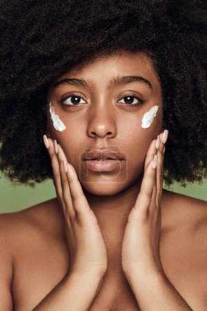 Photo for Young African American female with Afro hair and perfect smooth skin applying moisturizing cream on face looking at camera - Royalty Free Image