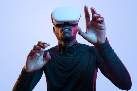 Foto de Atento joven afroamericano en traje negro tocando la pantalla invisible mientras explora el ciberespacio en gafas VR contemporáneas contra fondo azul - Imagen libre de derechos