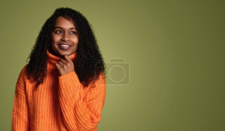 Téléchargez les photos : Une jeune femme souriante en pull orange confortable pose avec ses longs cheveux bouclés pour une séance photo portrait et cherche à espace vide en studio sur fond vert - en image libre de droit
