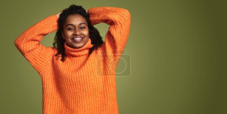 Téléchargez les photos : Femme afro-américaine joyeuse avec des cheveux bouclés portant un pull à col roulé orange vif et tenant les mains derrière la tête tout en regardant la caméra sur fond vert - en image libre de droit