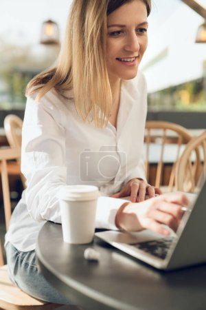 Téléchargez les photos : Cultiver jeune pigiste femme positive avec de longs cheveux blonds en chemise blanche souriant et tapant sur ordinateur portable, tout en étant assis sur la terrasse du café avec tasse de café pendant le travail à distance - en image libre de droit