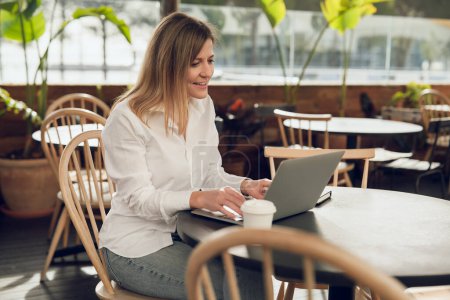 Photo for Delighted adult self employed woman with long brown hair in smart casual wear, smiling and working remotely on laptop while sitting at table in cafe with coffee cup - Royalty Free Image