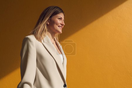 Téléchargez les photos : Vue latérale du jeune manager féminin ravi avec de longs cheveux blonds dans une tenue formelle élégante, souriant et regardant loin avec confiance tout en se tenant près du mur jaune vif sur la rue - en image libre de droit