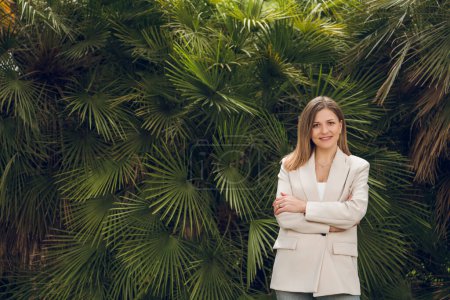 Téléchargez les photos : Jeune entrepreneure positive aux longs cheveux bruns en costume beige élégant, souriante et regardant la caméra tout en se tenant près de plantes tropicales vertes luxuriantes aux bras croisés - en image libre de droit