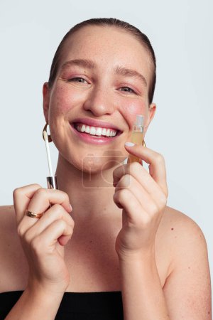 Foto de Sonriente modelo femenino joven demostrando producto cosmético brillo labial en estudio contra fondo gris mirando a la cámara - Imagen libre de derechos