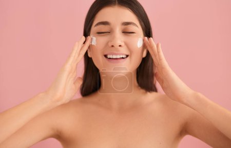 Téléchargez les photos : Joyeux jeune femelle aux épaules nues appliquant de la crème hydratante sur le visage avec les yeux fermés sur fond rose - en image libre de droit