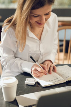 Téléchargez les photos : Crop souriant femme rédacteur prendre des notes dans le carnet tout en buvant café à emporter dans un café en plein air - en image libre de droit