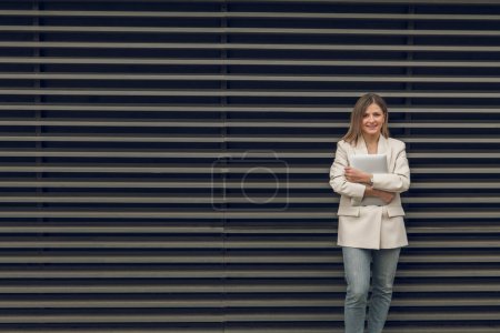 Téléchargez les photos : Jeune femme d'affaires positive en veste et jeans debout sur fond rayé avec ordinateur regardant la caméra - en image libre de droit