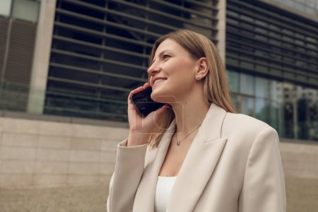 Photo for Low angle of positive adult female entrepreneur with long blond hair in elegant suit, smiling and looking away while talking on mobile phone on city street - Royalty Free Image