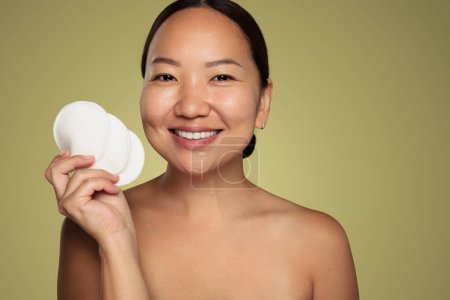 Photo for Cheerful young female with bare shoulders showing white makeup removal pads smiling looking at camera against green background - Royalty Free Image
