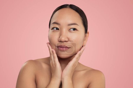 Photo for Young ethnic female with bare shoulders standing in studio after skincare routine looking away against pink background - Royalty Free Image