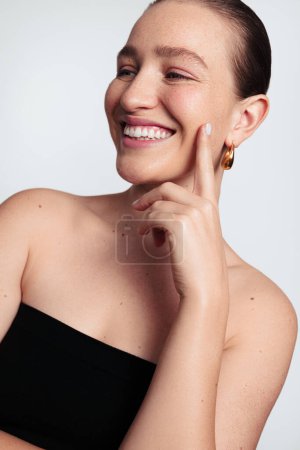 Foto de Feliz joven hembra con hombros desnudos sonriendo y tocando la barbilla en una sesión de fotos de belleza mientras está de pie sobre fondo blanco en el estudio - Imagen libre de derechos