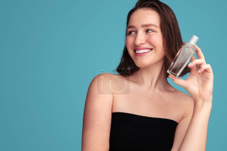 Foto de Alegre modelo femenina bastante joven con hombros desnudos sonriendo mientras muestra botella de vidrio de perfume fragante sobre fondo azul - Imagen libre de derechos